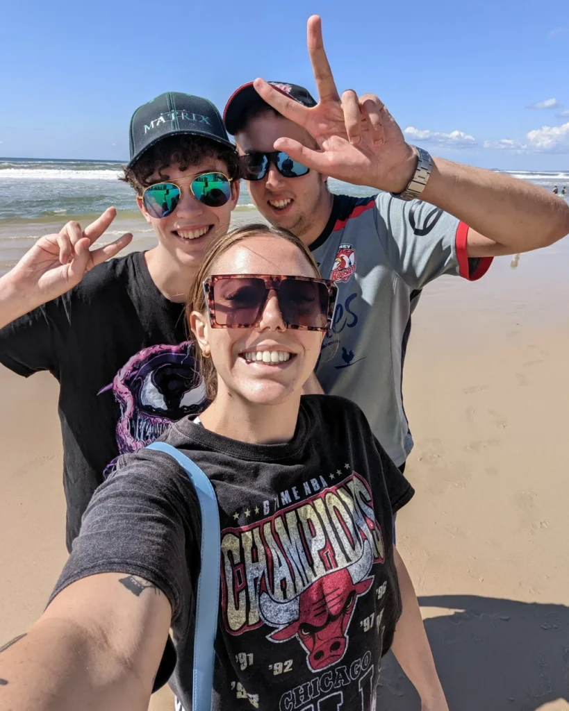 Brunette taking a selfie with two boys smiling and making peace signs in the back, on the beach